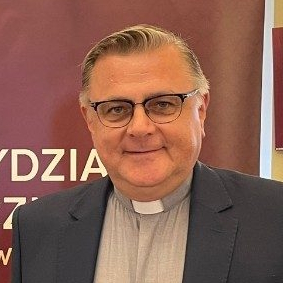 Uśmiechnięty ksiądz w okularach, z kolatką i w ciemnej marynarce.